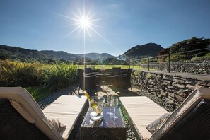 Un espacio al aire libre iluminado por el sol con una mesa para dos, con vistas a un paisaje escénico con colinas y un muro de piedra.