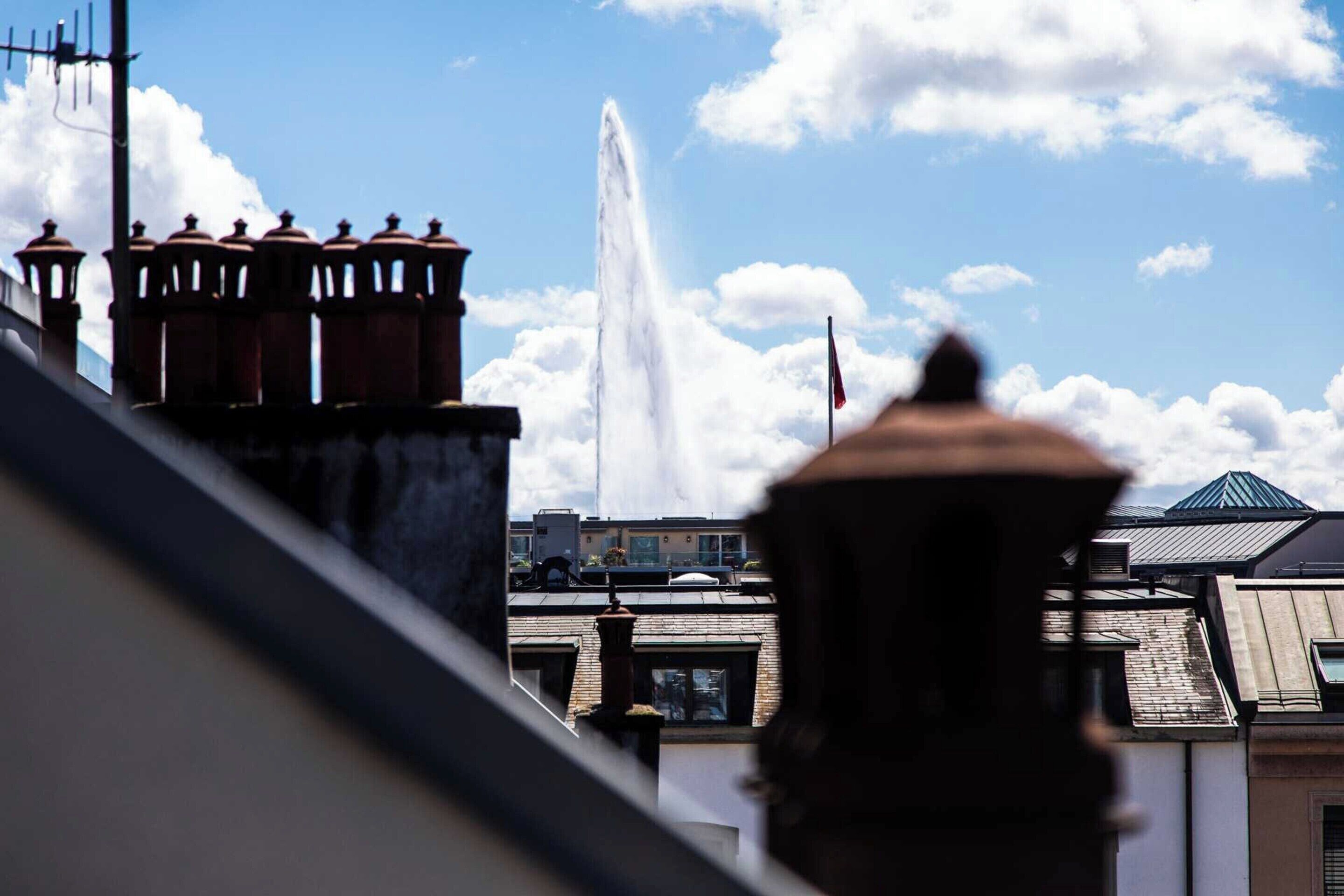 A building with a prominent water fountain in the background, surrounded by other structures.