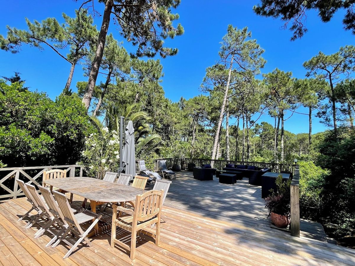 Une terrasse extérieure avec une table et des chaises en bois, entourée de grands arbres et surplombant un ciel bleu dégagé.