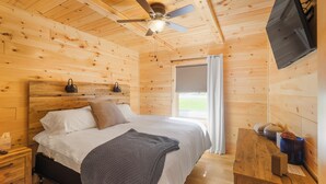 A wooden cabin bedroom with a bed, a ceiling fan, a TV mounted on the wall, and a window with curtains. - Seasons on Oak Short walk to best Fly Fishing in Northeast Cozy Cabin Deposit NY (Deposit)