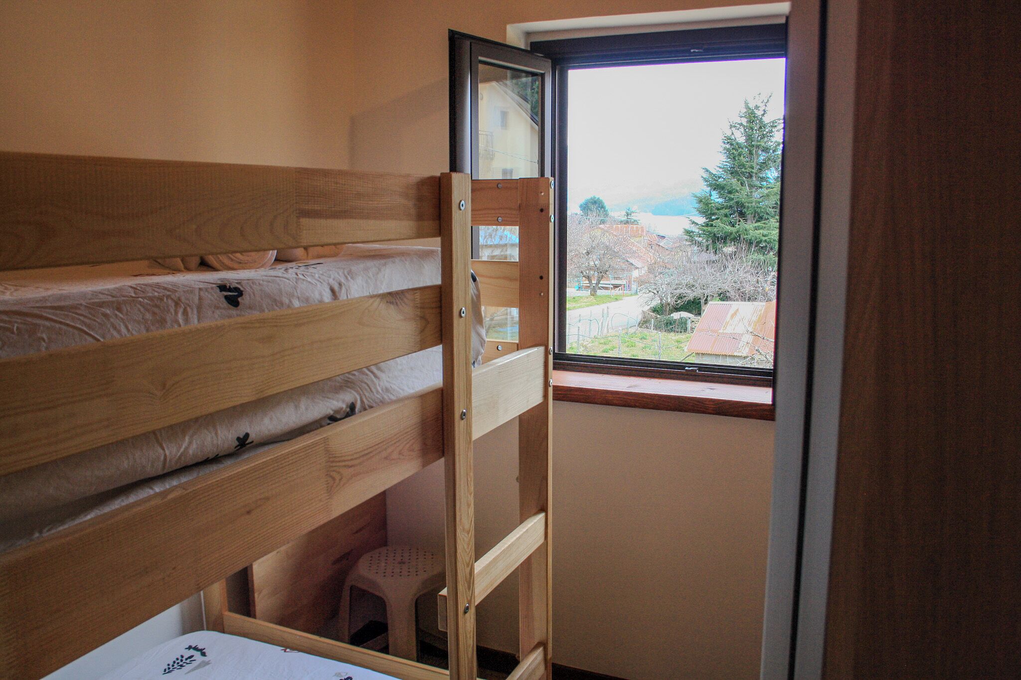 A bunk bed with a wooden frame and a small stool underneath, a window with a view of houses and trees, and a door frame.