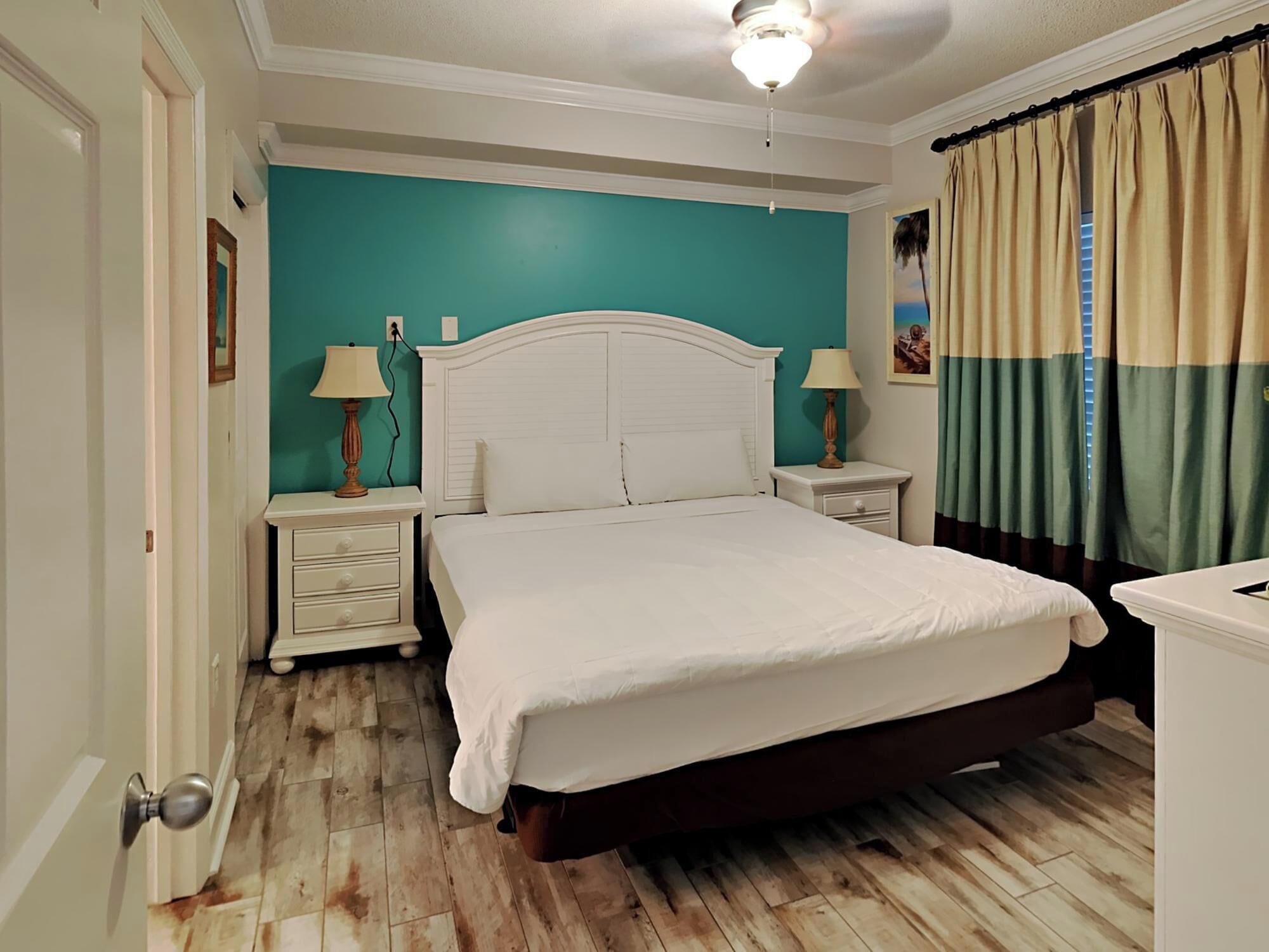 A bedroom with a white bed, teal walls, wooden flooring, and a ceiling fan.