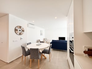 A modern living room with a round dining table, grey patterned chairs, a blue sofa, a flat-screen TV, and a wall-mounted clock.