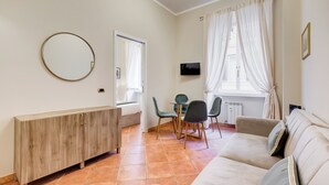 Un salon comprenant un canapé, une table à manger avec des chaises, un téléviseur fixé au mur et une fenêtre avec des rideaux.