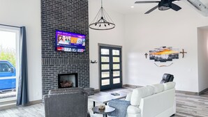 A modern living room with a fireplace, a television mounted on a brick wall, a ceiling fan, and a white sofa. - WhiteTail Cabin with Shuffleboard, Fireplace, WiFi, South of Norman (Purcell)