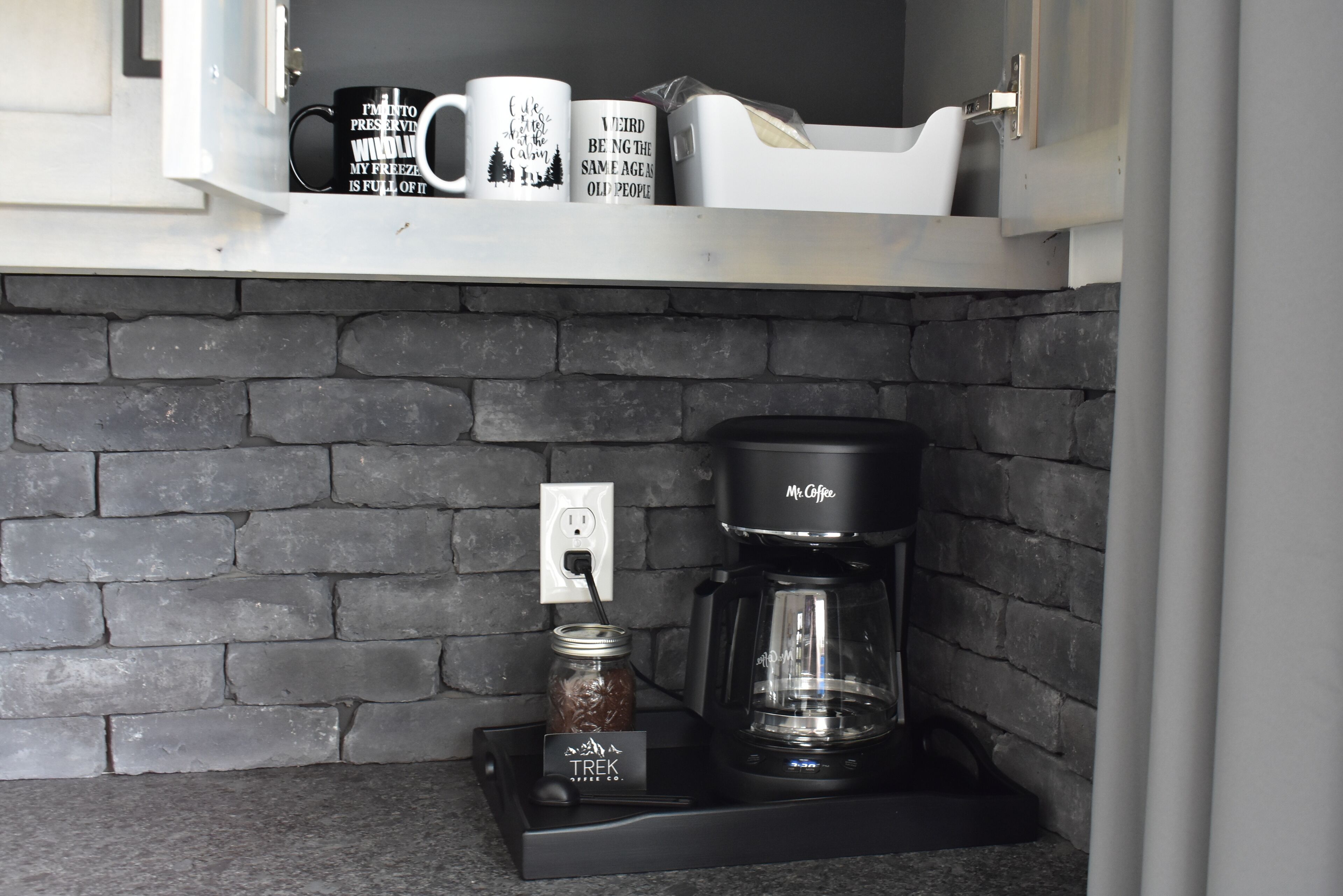 A kitchen area with a coffee maker, a jar of coffee, and two mugs on a shelf.
