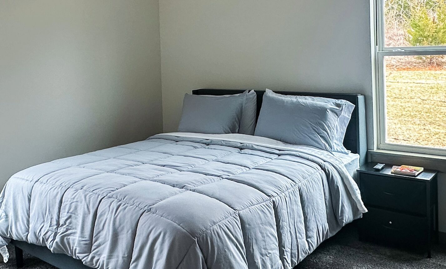 A bedroom with a bed, two pillows, a nightstand, and a window with a view of the outdoors.