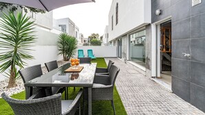 Un área de comedor exterior moderna con una mesa de vidrio, sillas de mimbre y una sombrilla grande.
