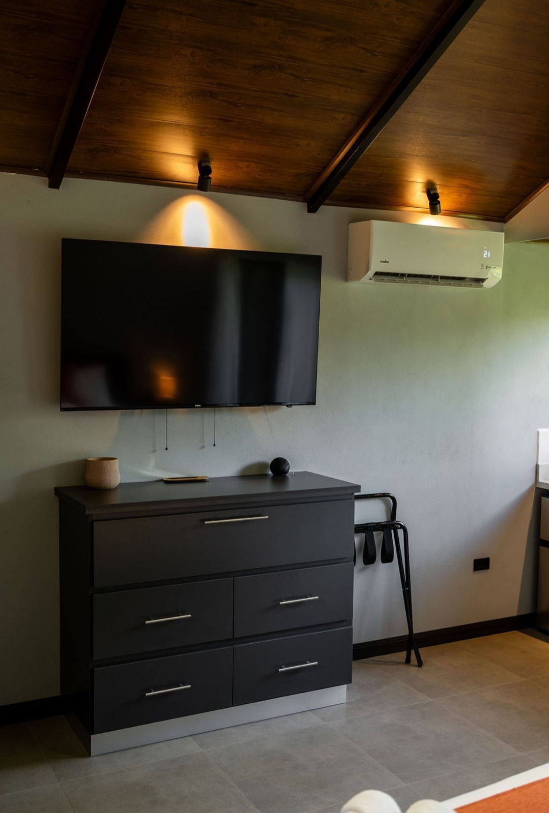 A modern room with a large flat-screen TV mounted on the wall, a dark wooden cabinet with drawers, and an air conditioning unit.