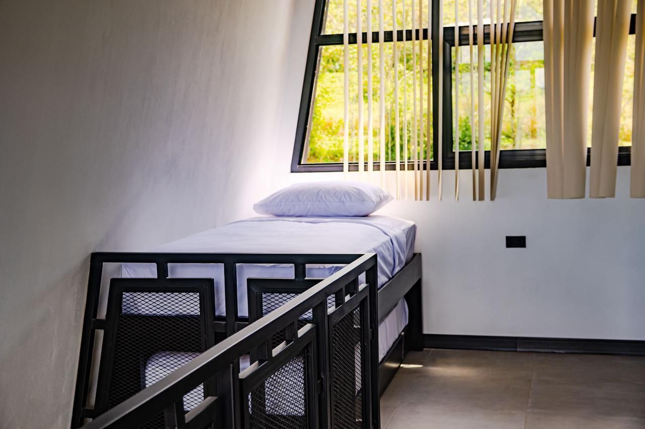 Un lit simple avec un oreiller blanc et un cadre en métal noir, une fenêtre avec des stores verticaux et une vue sur la verdure à l’extérieur.