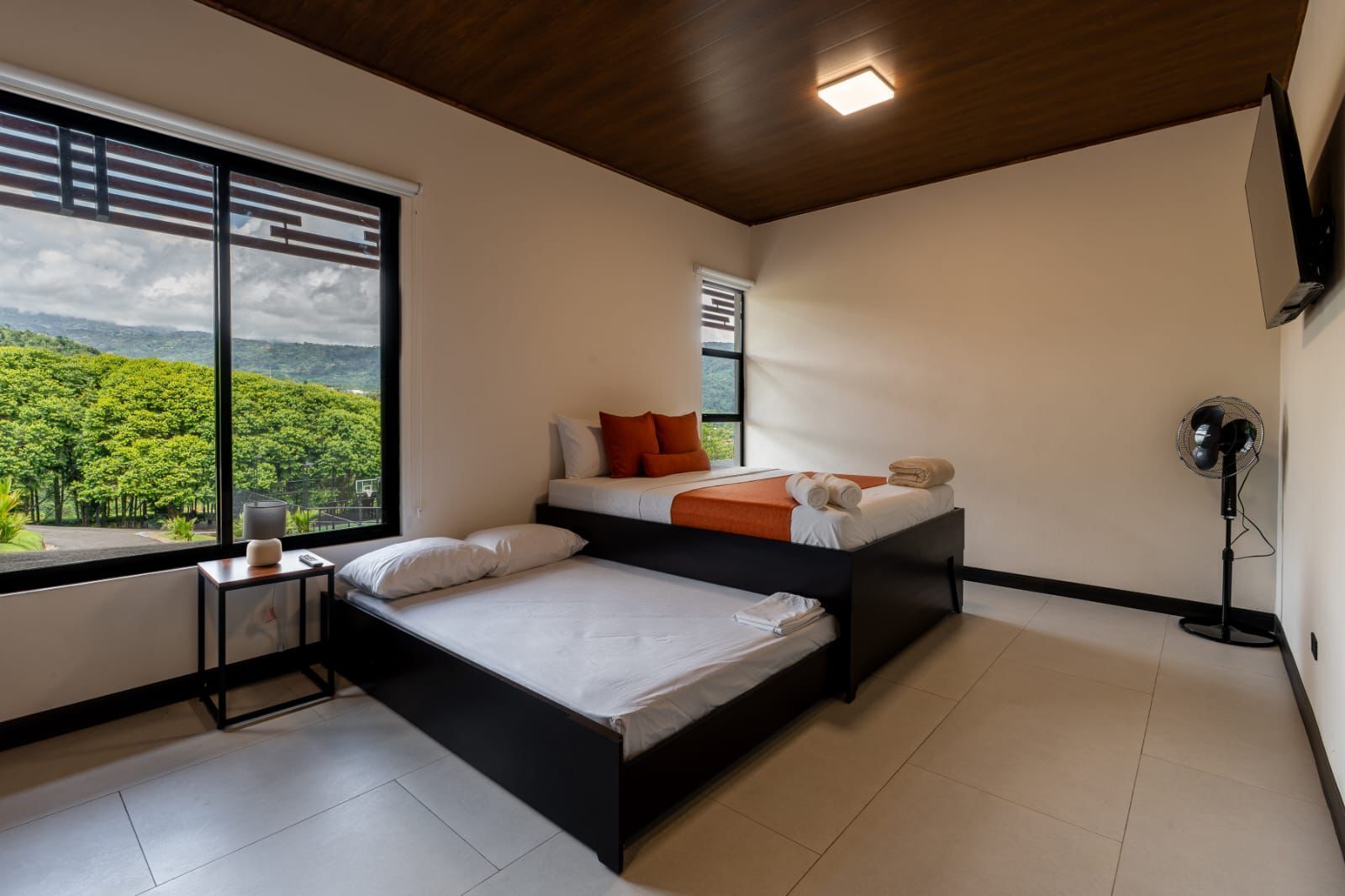A modern bedroom with a large window, a bed with white and orange bedding, a bedside table with a lamp, and a standing fan.