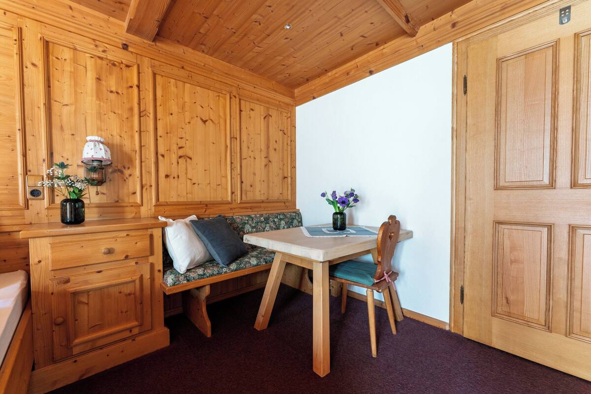 Een houten blokhut van binnen, met een houten bank, een tafel met een bloevas en een deur.