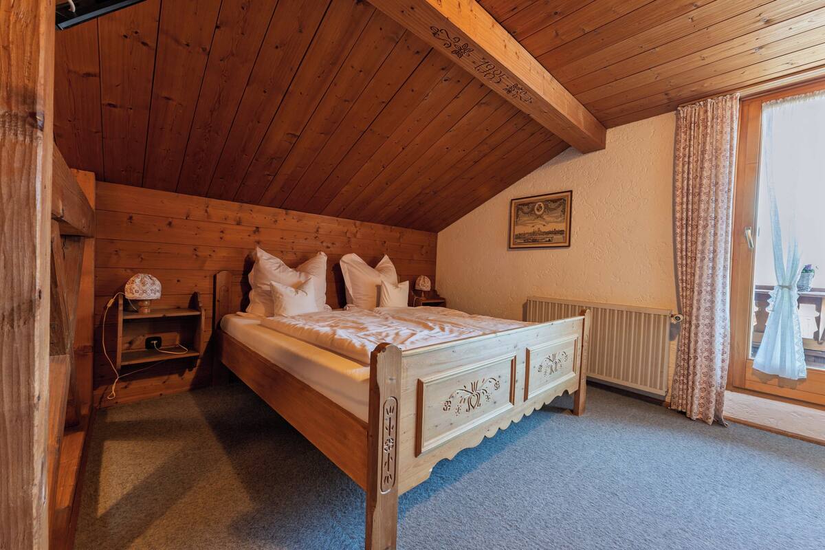 Een kamer met houten frame, voorzien van een bed, nachtkastje, lamp en raam met gordijnen.