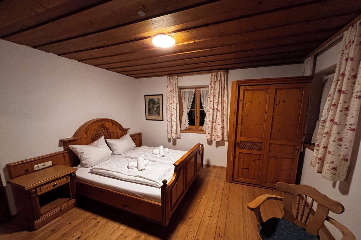 Een bed met houten frame en witte lakens, een houten nachtkastje, een houten stoel, een houten garderobe en een raam met bloemen gordijnen.