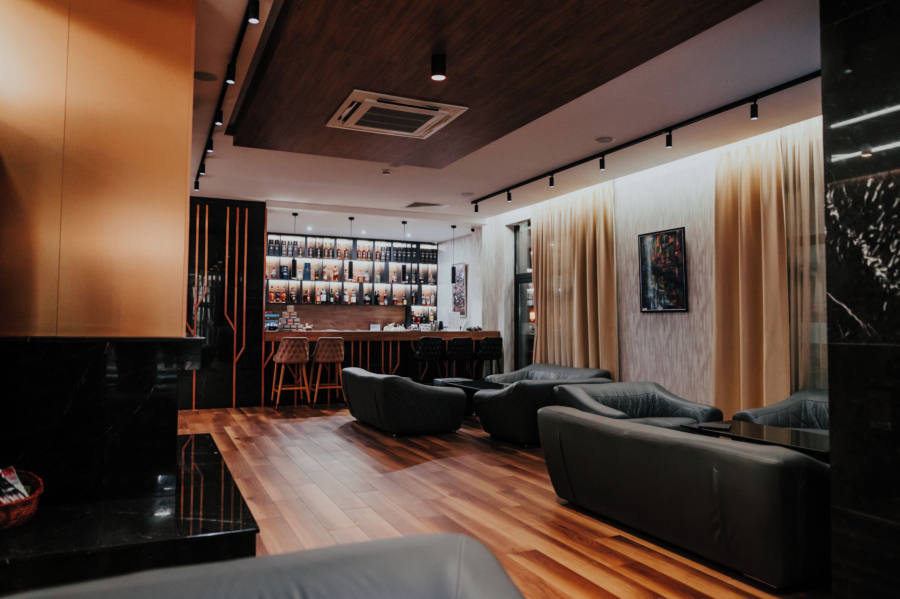 A modern bar area with a wooden floor, black leather seating, and a bar stocked with various bottles.