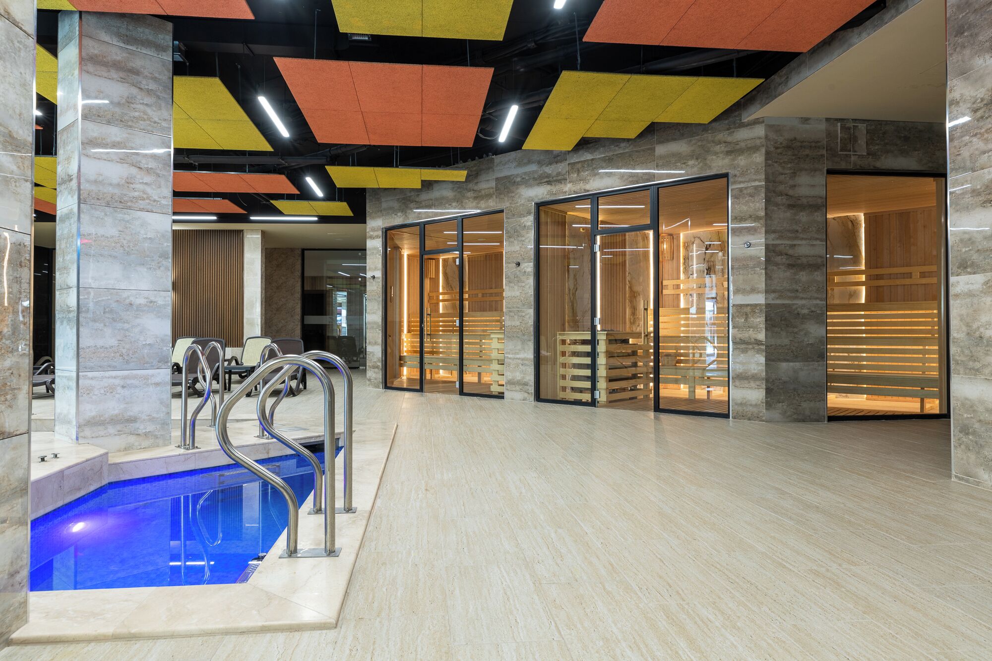 Zona d'una piscina coberta moderna, amb parets de vidre transparent, una piscina blava i un sostre amb motius geomètrics.