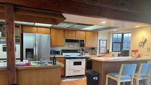 A kitchen with wooden cabinets, a stainless steel refrigerator, a microwave, a stove, and a sink. There are two bar stools and a trash can.