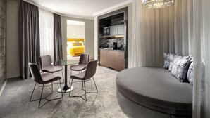 Een moderne hotelkamer met een ronde zithoek, een eettafel met stoelen, een keukenhoekje en een bed met een geel hoofdeinde.