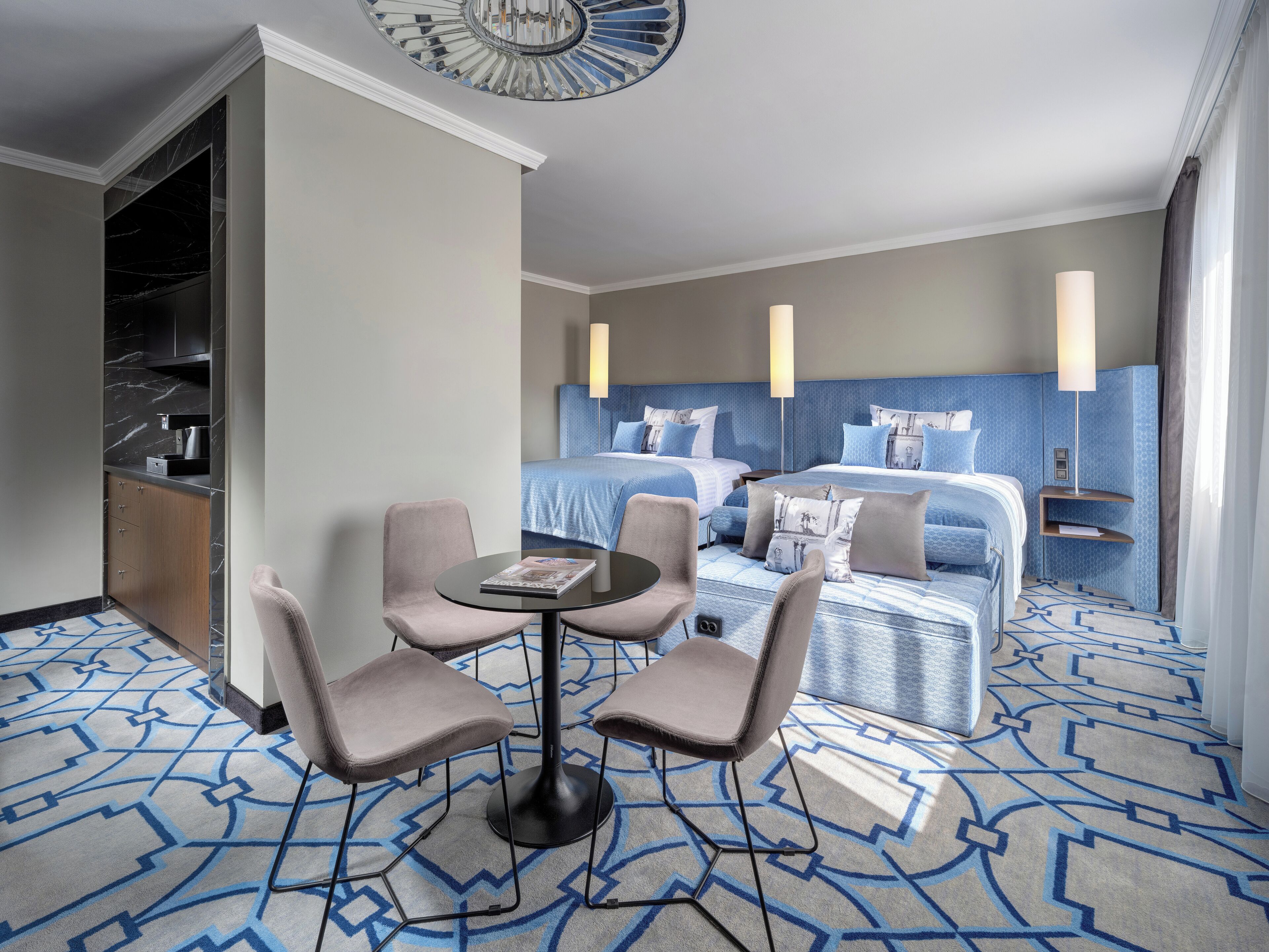 A hotel room with a bed, two bedside tables, a desk with chairs, a round table, and a patterned carpet.
