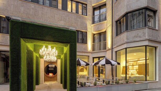 Een hotelentree met een groene haagboog, een kroonluchter en terrasmeubels.