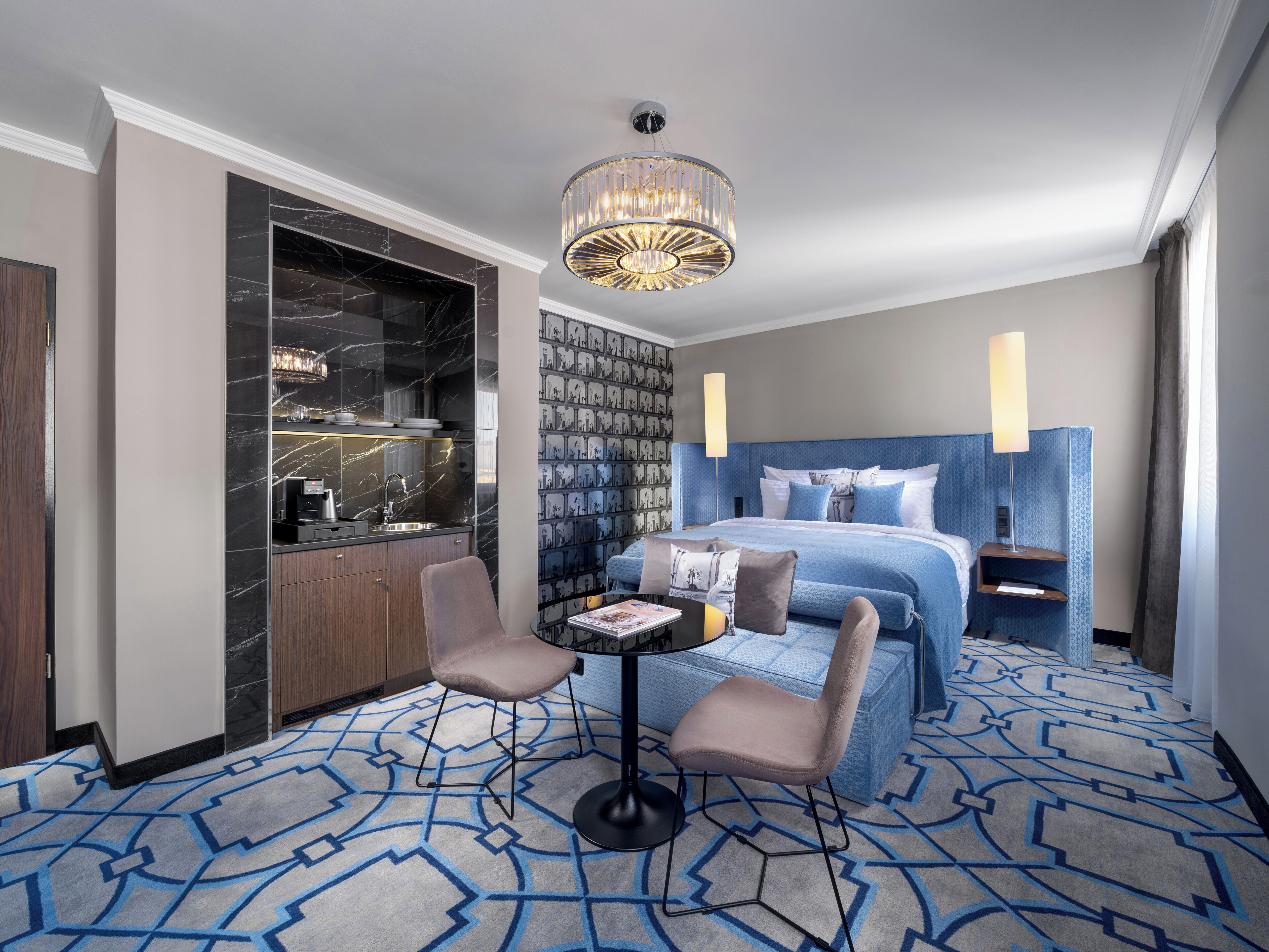 A hotel room with a marble bathroom, a patterned carpet, a bed with blue bedding, a small table with a magazine, and a modern chandelier.