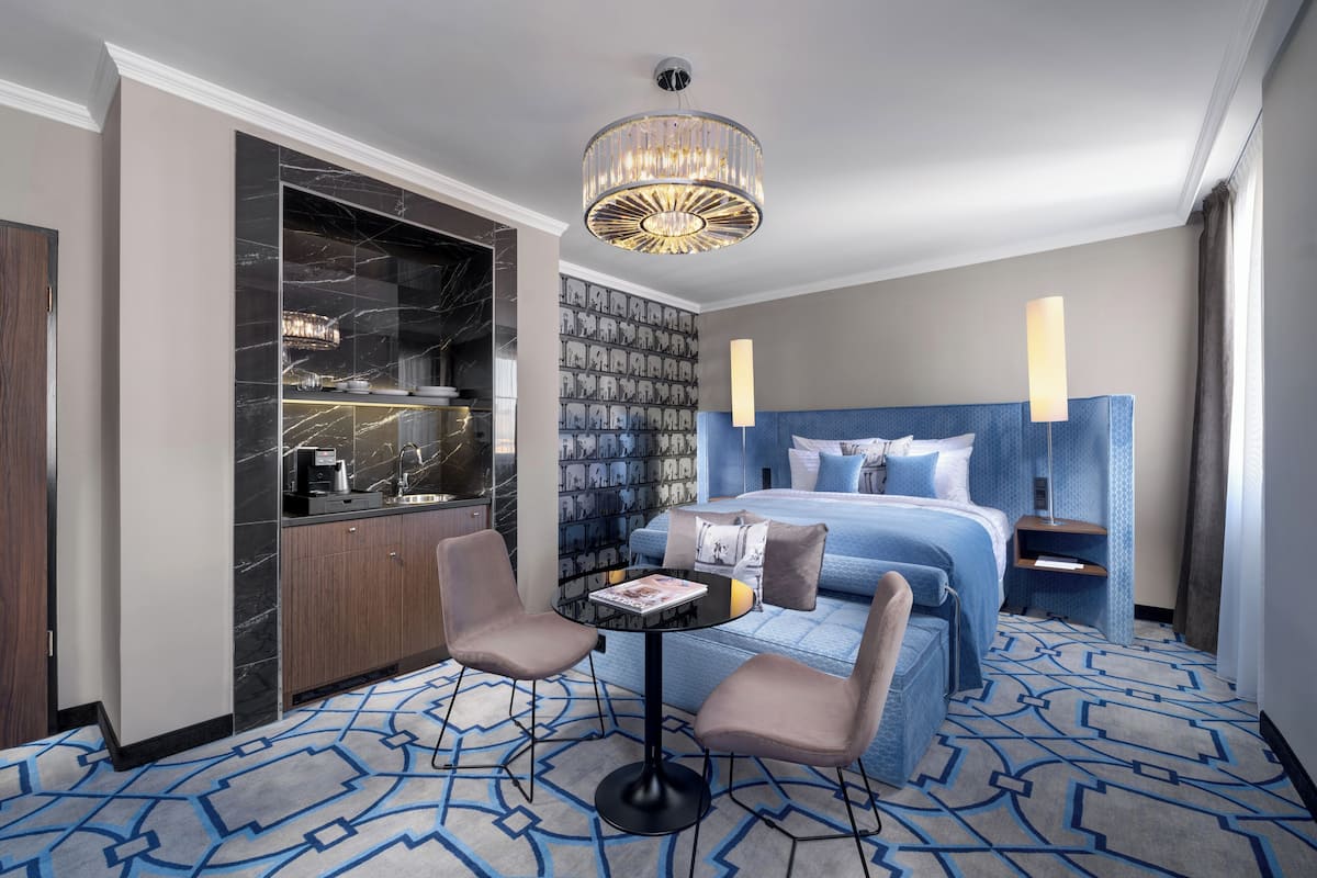 A hotel room with a marble bathroom, a patterned carpet, a bed with blue bedding, a small table with a magazine, and a modern chandelier.