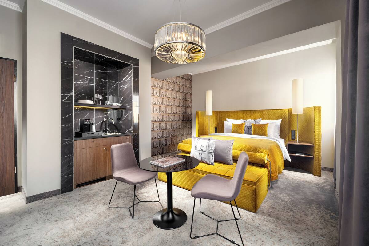 A modern hotel room with a marble kitchen, a large bed with a yellow headboard, two chairs around a small table, and a chandelier.