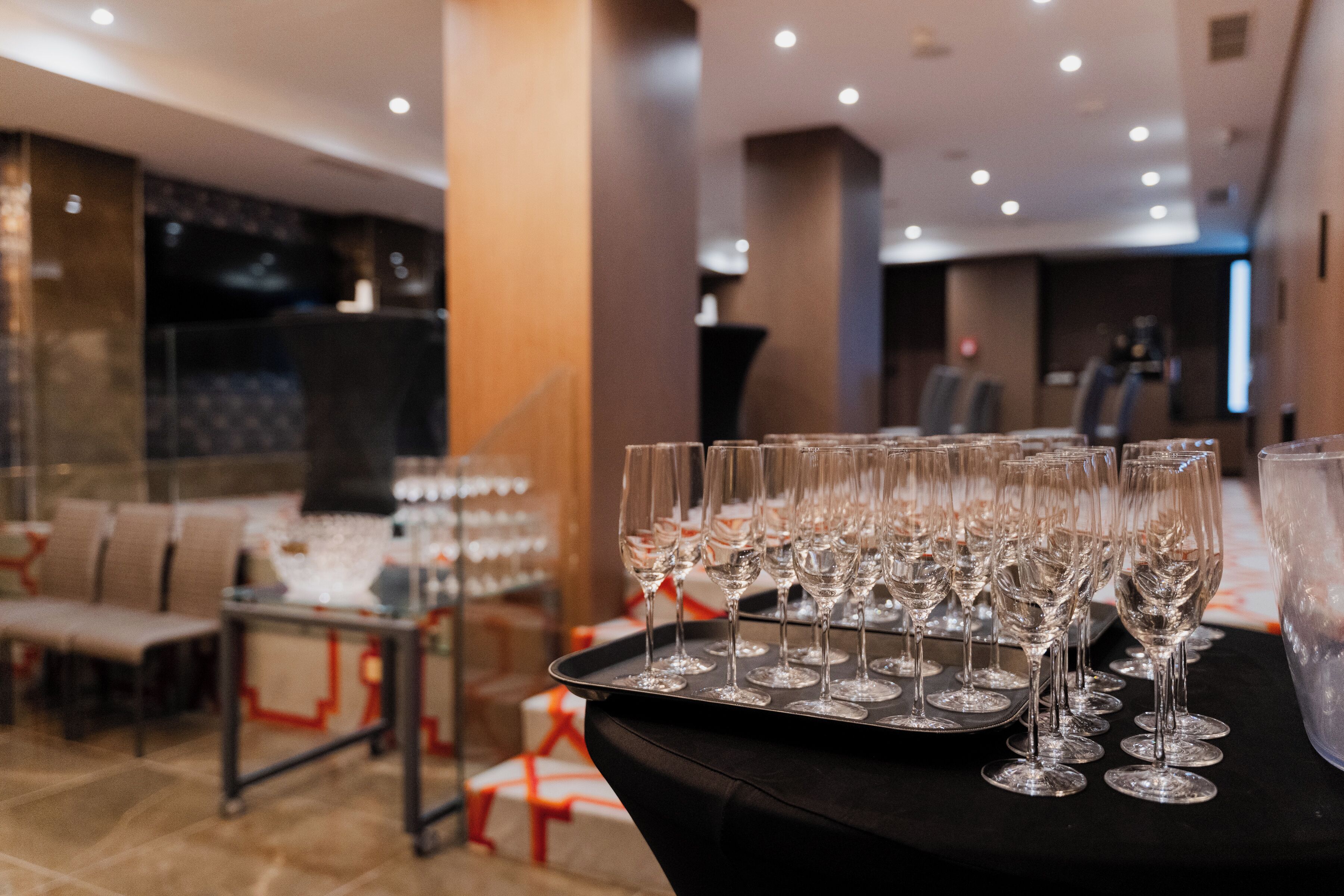 A table with multiple wine glasses arranged on a tray, a seating area with chairs, and a reception desk in a modern hotel lobby.