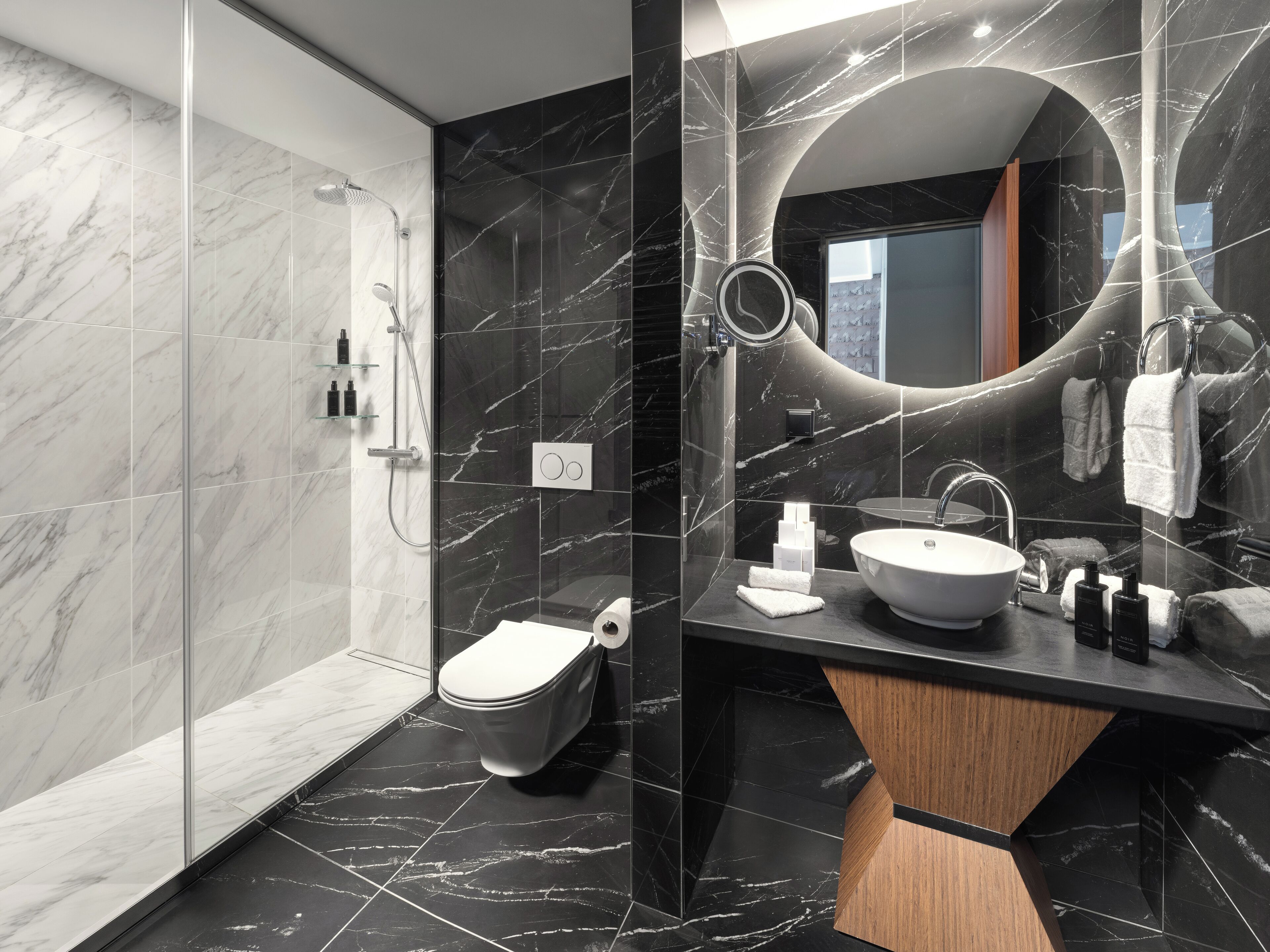 Un baño moderno con ducha envuelta en cristal, inodoro, lavamanos con tocador de madera y un espejo grande.