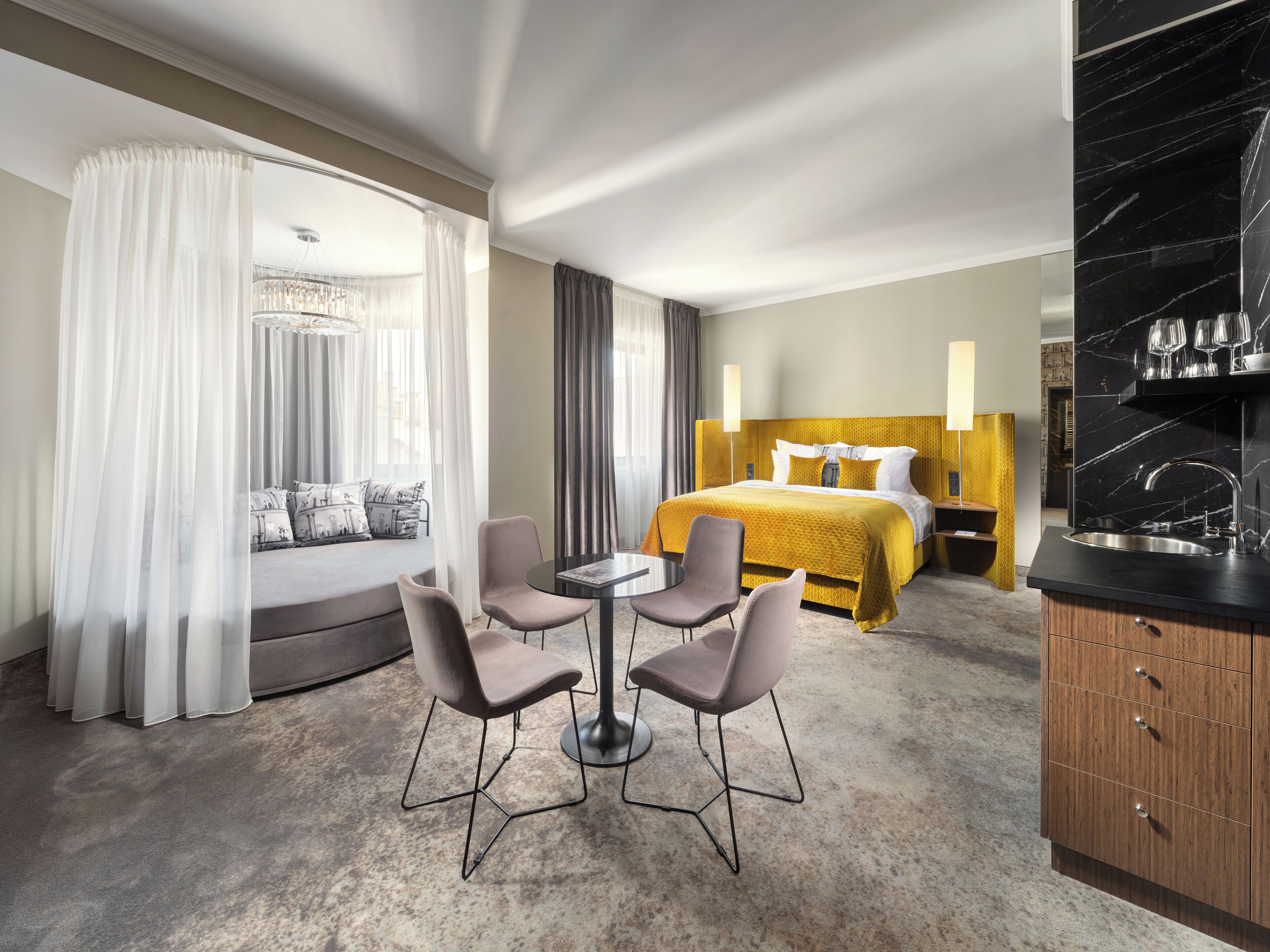 A hotel room with a bed, a dining table with four chairs, a sofa, a large window with sheer curtains, and a kitchen area with a black marble countertop.