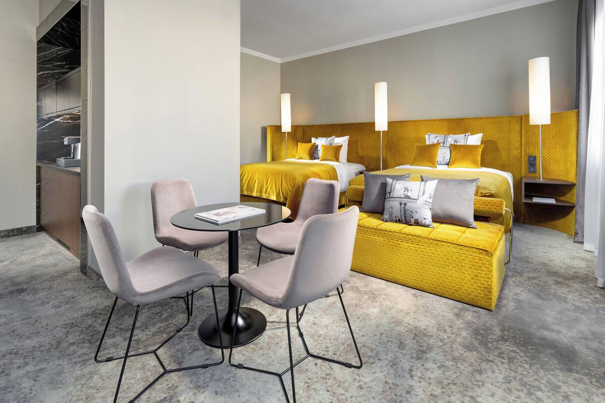 A hotel room with a bed, two bedside tables, a round table with four chairs, and a small kitchenette.