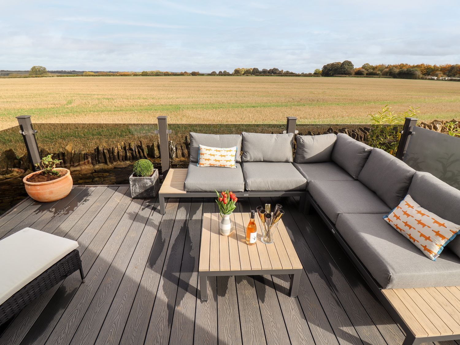 An outdoor seating area with a wooden deck, grey cushioned sofa, and a small table with drinks and flowers.
