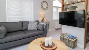 Una sala de estar con un sofá gris, una mesa redonda de madera con velas, un televisor de pantalla plana y una estantería con objetos decorativos.