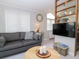 Una sala de estar con un sofá gris, una mesa redonda de madera con velas, un televisor de pantalla plana y una estantería con objetos decorativos.