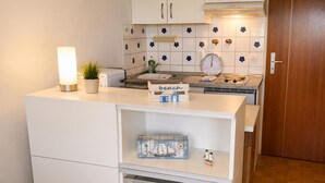 Een compacte keuken met witte kastjes, een betegelde spatwand, een klok en een klein plankje met een nautisch decoratie.