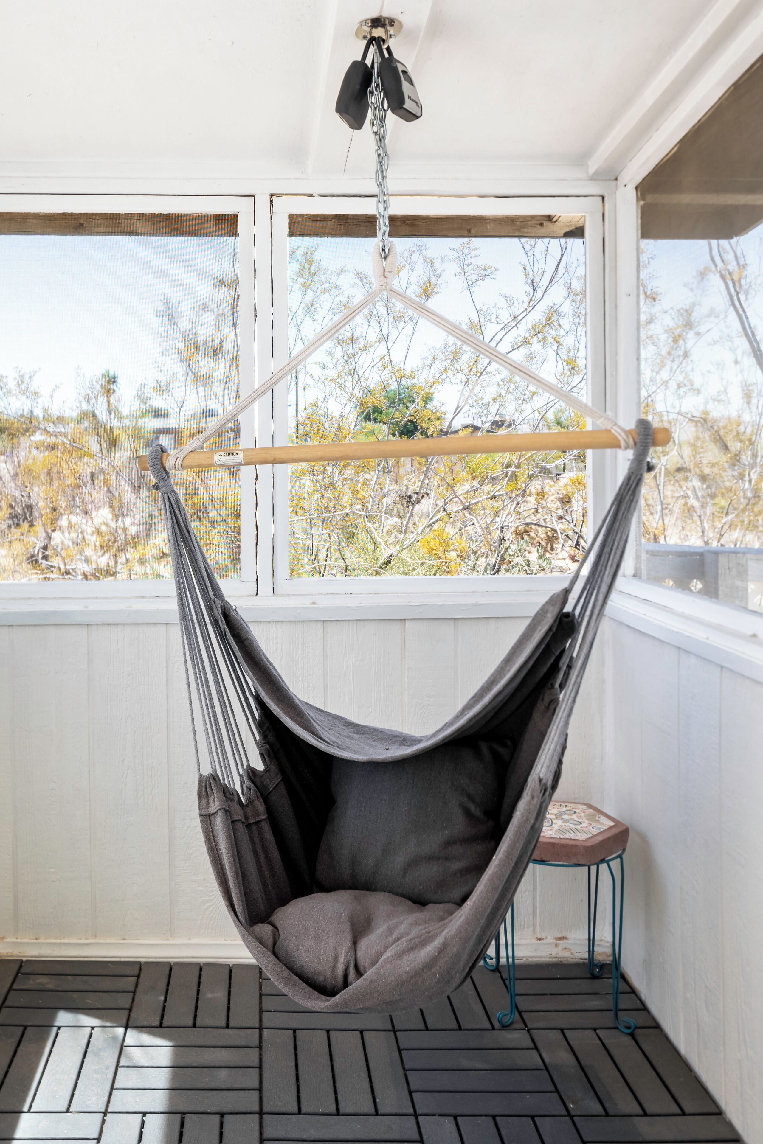 Una silla hamaca colgada de una cadena en el techo, un taburete pequeño y vistas a árboles a través de una ventana.