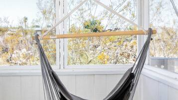Una silla hamaca colgada de una cadena en el techo, un taburete pequeño y vistas a árboles a través de una ventana.