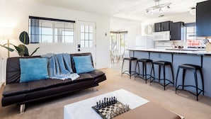 A modern living room with a black leather sofa, a coffee table with a chess set, and a kitchen area with bar stools.