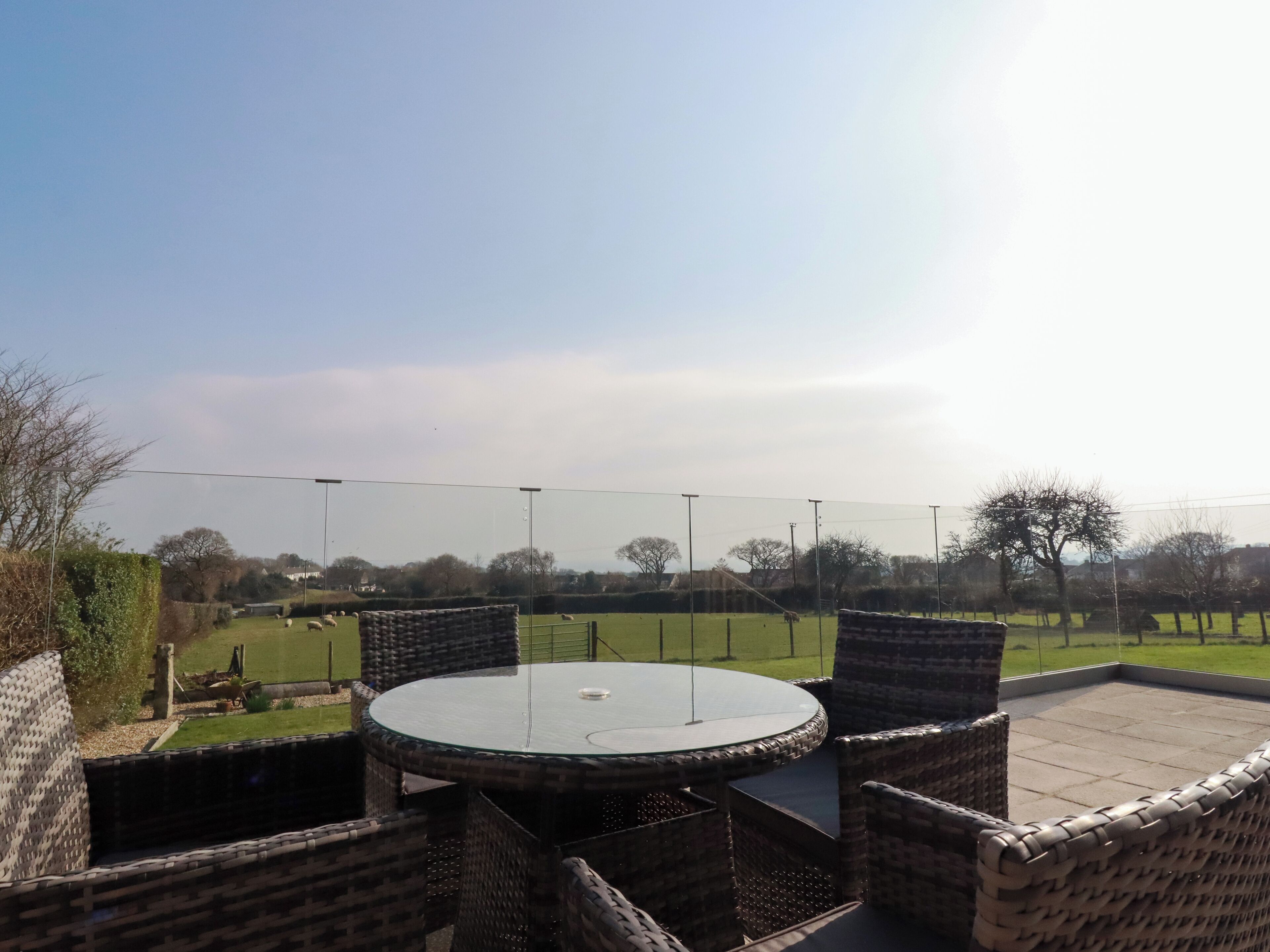 A round outdoor table with wicker chairs, surrounded by a grassy area with a fence and trees in the background.