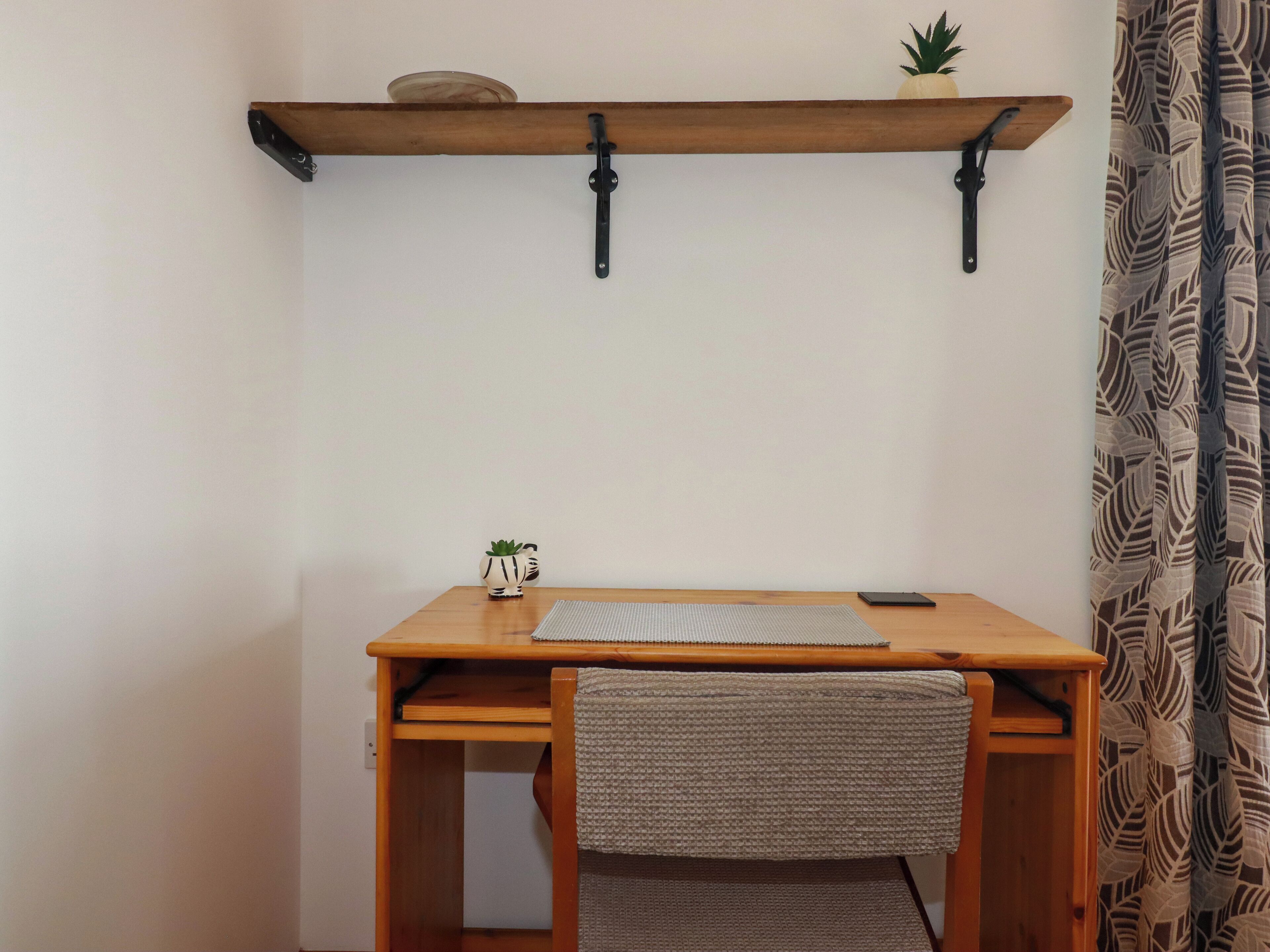 A wooden desk with a woven chair, a small potted plant, and a smartphone on the desk.
