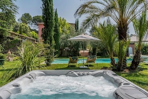 Ein Whirlpool, umgeben von einem üppigen Garten mit Pool und Liegestühlen.