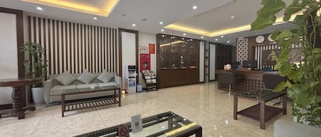 A modern hotel lobby with a sofa, a coffee table, a reception desk, and a large plant.