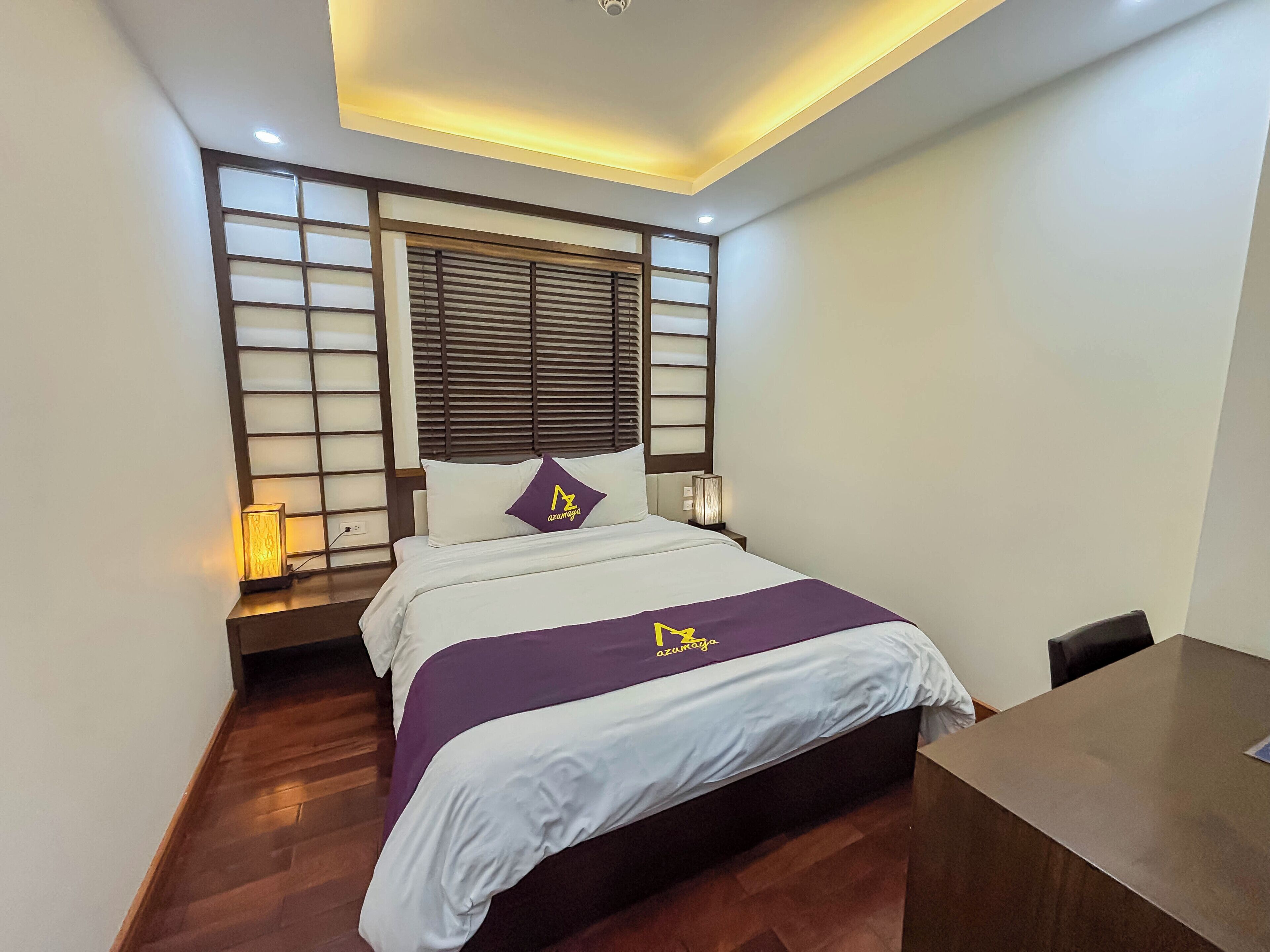 Une chambre d’hôtel moderne équipée d’un lit, de tables de chevet, d’un bureau et d’une fenêtre avec des stores.