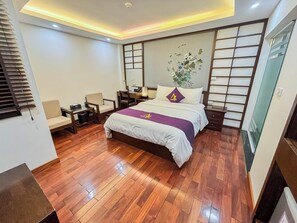 A modern hotel room with a wooden floor, a bed with a white and purple bedspread, a desk with a chair, a nightstand, and a large window with blinds. - Azumaya Hotel Dao Tan (Hanoi)