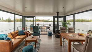A spacious indoor area with a large window overlooking a body of water, a wooden table, a sofa, and a grill. - Hausboot am Prerauer Stich in Zehdenick (Zehdenick)