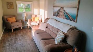 Un salon avec un canapé beige, un fauteuil marron, une petite table d’appoint avec une lampe, et un tableau représentant une jetée accroché au mur.