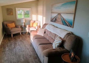 Un salon avec un canapé beige, un fauteuil marron, une petite table d’appoint avec une lampe, et un tableau représentant une jetée accroché au mur.