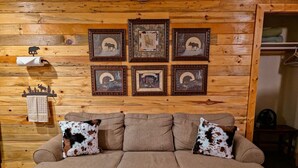 A brown sofa with two cowhide-patterned pillows, a wooden wall with six framed pictures, and a small shelf with a towel and a fan. - Cozy Cabin 1 Block to Lake/In Town/Hummingbird (Grand Lake)