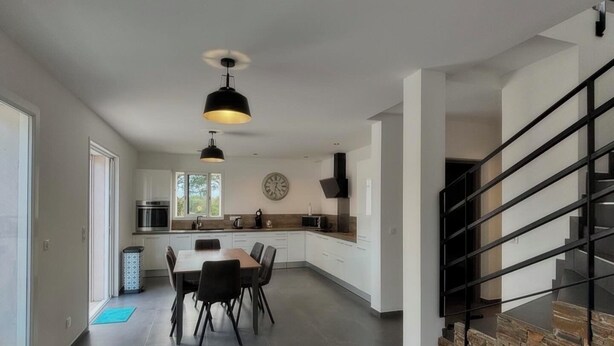 A spacious modern kitchen with a dining area, a clock on the wall, and a staircase with a stone accent wall. - Villa Melendria, Ghisonnaccia (Ghisonaccia)