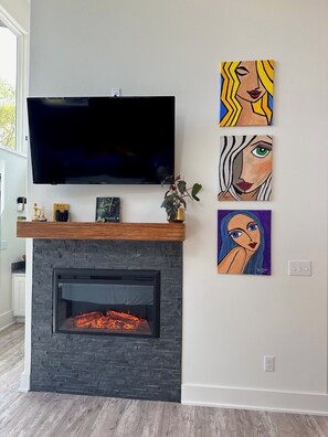 A living room with a flat-screen TV mounted above a stone fireplace, three paintings on the wall, and a small plant on the mantel.